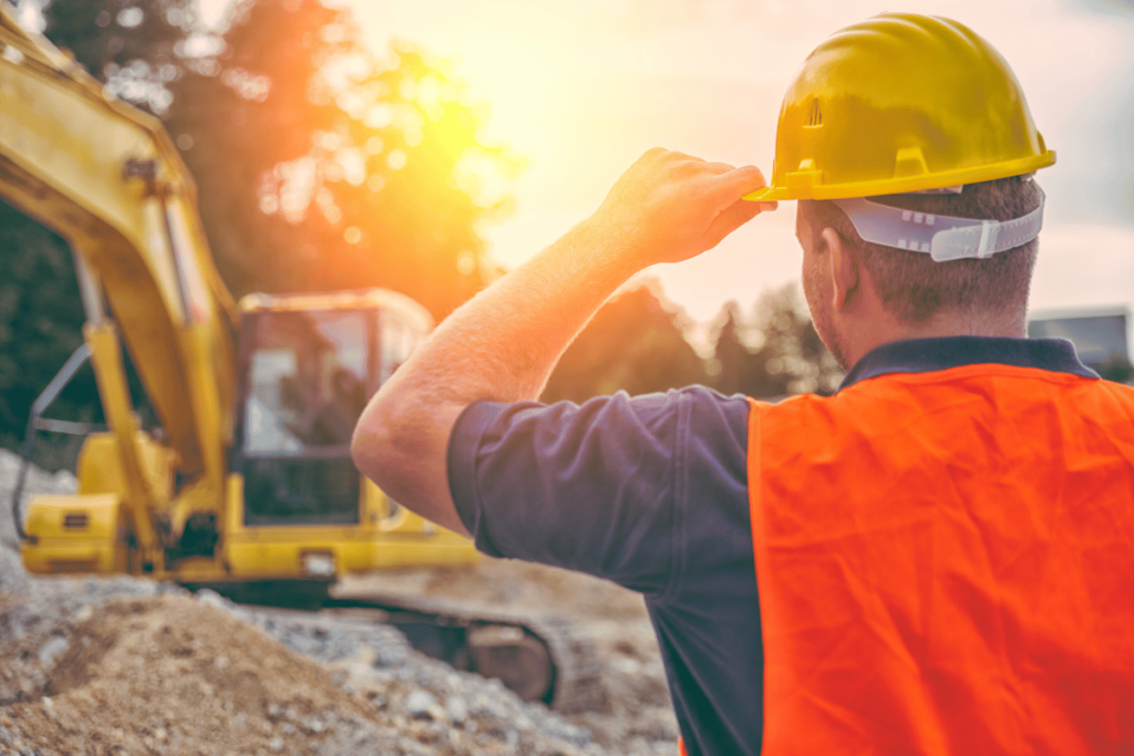 Lavoratore edile con casco giallo e giubbotto arancione supervisiona un cantiere al tramonto.